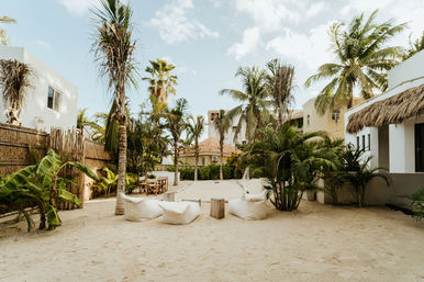 Sandy tropical courtyard with palm trees, white beanbag loungers and wooden dining table set between low coastal villas under a sunny blue sky