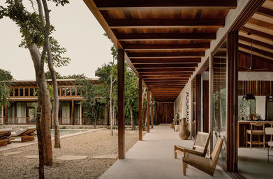 Serene tropical villa walkway with a long wooden pergola, sliding glass doors, lounge chairs and a pool framed by trees