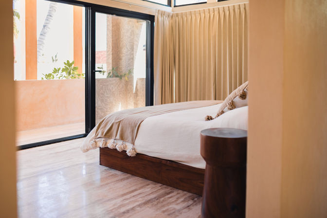 Sunlit coastal-style bedroom with wooden platform bed, cream linens and tassel throw, beige curtains and black-framed sliding glass doors opening to a balcony.