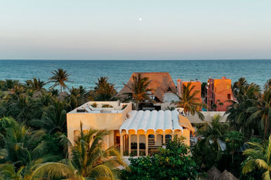 Aerial view of sunlit beachfront villas and thatched palapa nestled among tropical palm trees, turquoise ocean horizon at dusk with a pale moon rising above.