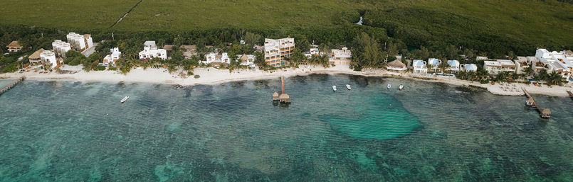 Aerial view of a tropical coastline with white-sand beach, turquoise shallow reef, beachfront villas, a wooden pier with thatched palapa, and small boats anchored offshore.