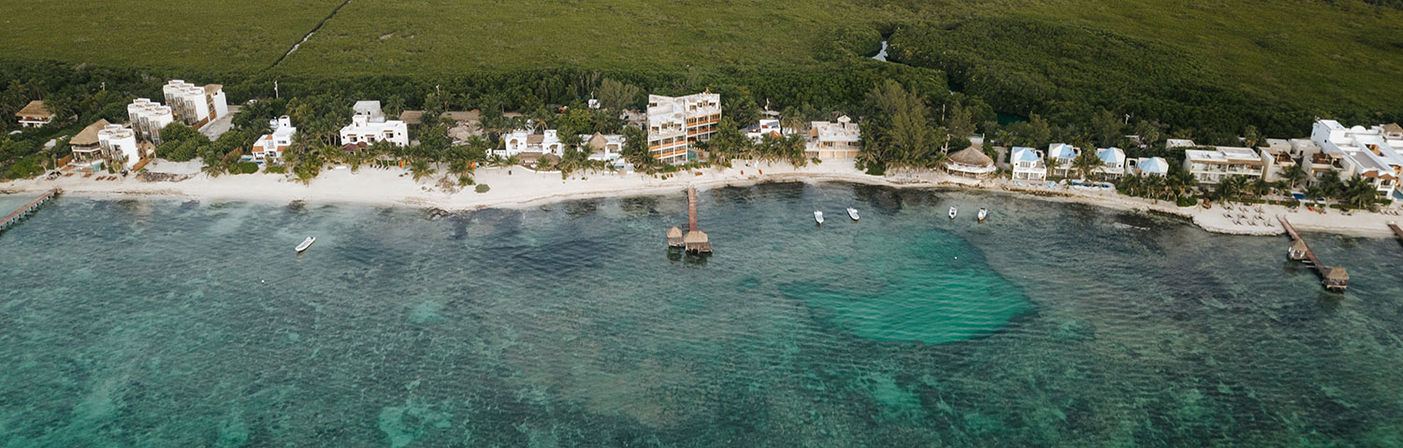Aerial view of a tropical coastline with white-sand beach, turquoise shallow reef, beachfront villas, a wooden pier with thatched palapa, and small boats anchored offshore.