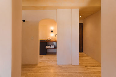 Warm minimalist hallway with arched alcove sink, soft wall sconce, wooden shelf of rolled white towels and travertine floor