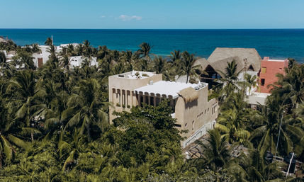 Aerial view of a sunlit beige stucco beachfront villa with arched windows and thatched palapa roofs nestled in dense palm trees along a turquoise ocean under a clear blue sky