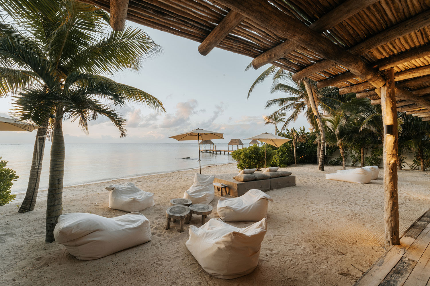 Tropical beachfront lounge under a wooden pergola at golden hour — palm trees, white sand, beanbag chairs and umbrellas with a wooden pier stretching into calm ocean
