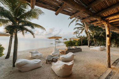 Tropical beachfront lounge under a wooden pergola at golden hour — palm trees, white sand, beanbag chairs and umbrellas with a wooden pier stretching into calm ocean