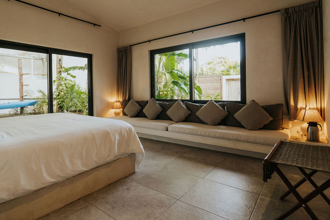 Cozy tropical villa bedroom with large bed, long cushioned sofa and neutral throw pillows, sliding glass door and windows opening to a lush garden, warm bedside lamps and soft natural light.