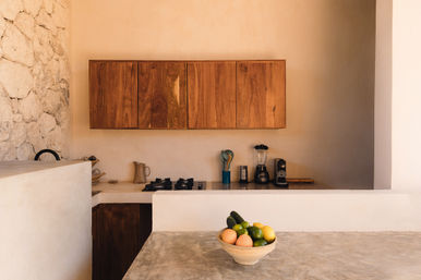 Sunlit minimalist Mediterranean-style kitchen with rough stone wall, warm wood cabinets, neutral plaster walls, concrete countertop, gas stove, blender and coffee maker on the counter, bowl of citrus and avocados in the foreground.