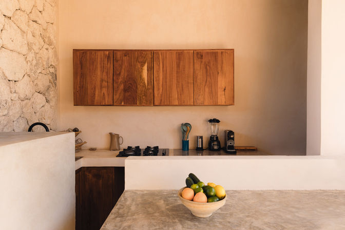Sunlit minimalist Mediterranean-style kitchen with rough stone wall, warm wood cabinets, neutral plaster walls, concrete countertop, gas stove, blender and coffee maker on the counter, bowl of citrus and avocados in the foreground.