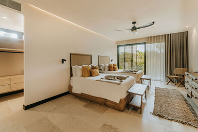 Sunlit modern coastal resort bedroom with two beds, wooden frames and benches, textured pillows and rug, ceiling fan, and sliding glass doors to tropical greenery