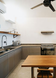 Modern minimalist urban apartment kitchen with L-shaped gray cabinets, wooden table and chair, stainless spring-coil faucet, microwave, gas range, range hood and ceiling fan.