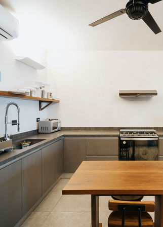 Modern minimalist urban apartment kitchen with L-shaped gray cabinets, wooden table and chair, stainless spring-coil faucet, microwave, gas range, range hood and ceiling fan.