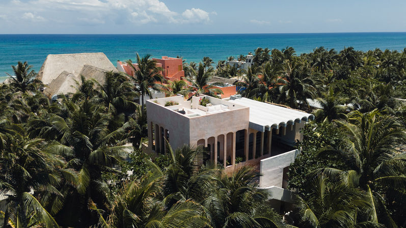 Aerial view of a modern beachfront villa with arched windows nestled among dense palm trees and colorful coastal homes, turquoise ocean on the horizon.