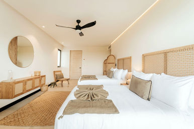 Bright, sunlit minimalist hotel bedroom with two beds dressed in white linens and tan throws, woven rattan headboards, wooden console with round mirror, black ceiling fan and natural jute rug.