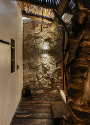 Cozy rustic rain shower with overhead black showerhead on a textured stone wall, warm accent lighting, slatted wooden roof, dark wooden plank floor and small wooden stool—outdoor-style bathroom vibe