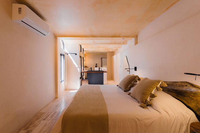 Sunlit minimalist bedroom with neutral plaster walls, king bed in white linens and beige tassel pillows, rustic wooden headboard, wall-mounted AC, arched window, and a small open kitchenette visible in the background.