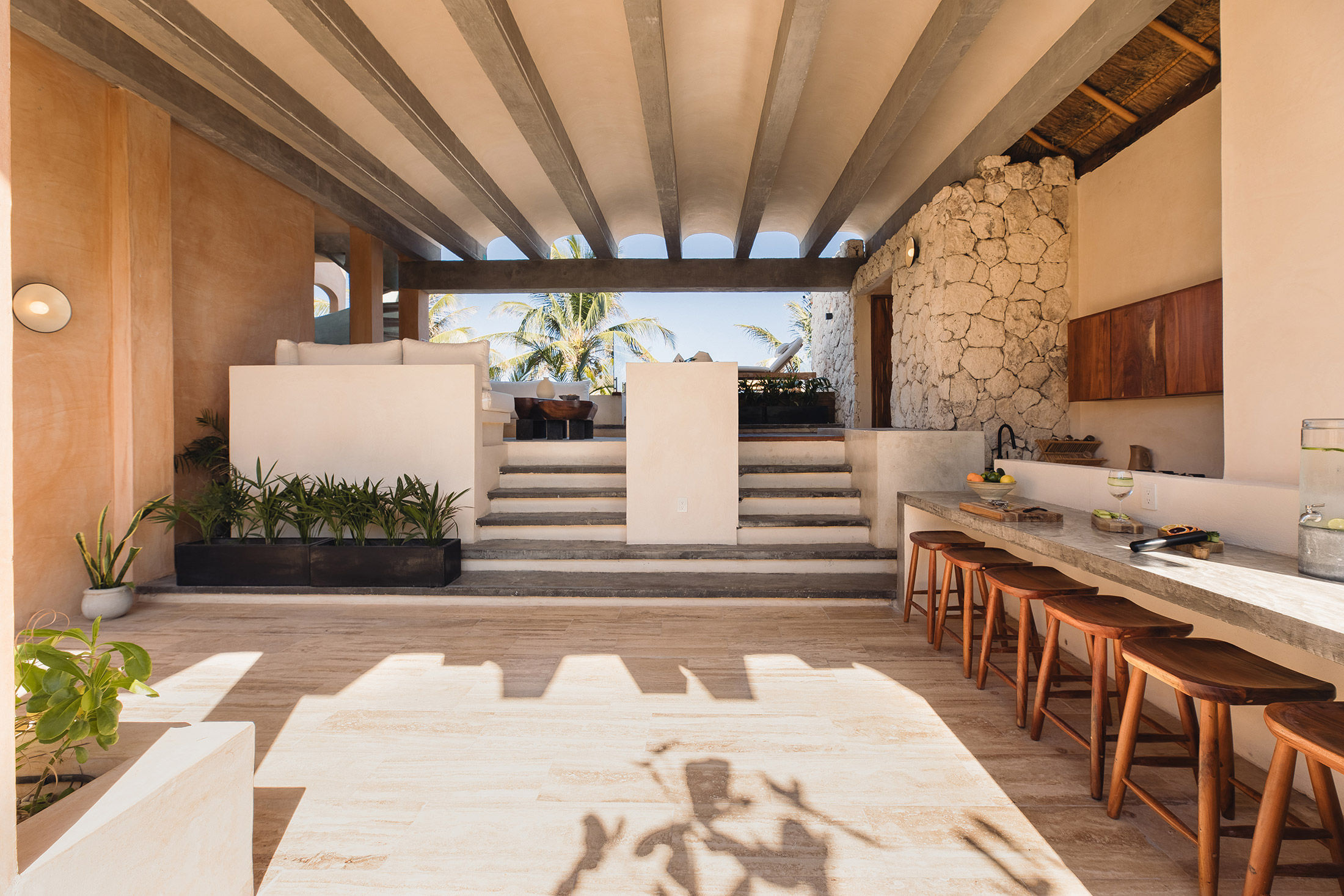 Sunlit open-air coastal villa living area and outdoor kitchen with exposed concrete beams, textured stone wall, raised white-sofa lounge, long stone breakfast bar with wooden stools, potted plants and palm trees visible beyond.