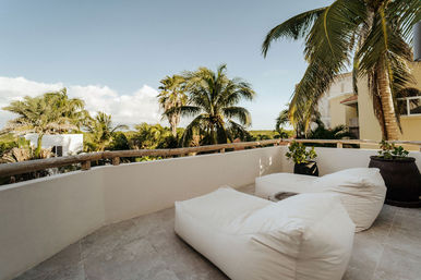 Sunny tropical balcony patio with two white beanbag lounge chairs, wooden railing, potted plants and palm trees under a clear blue sky — relaxing coastal vacation vibe.