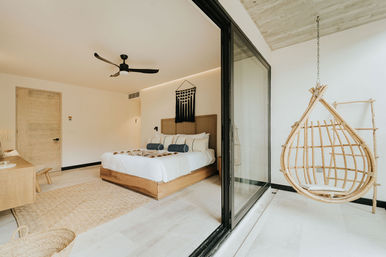 Sunlit modern minimalist bedroom with wooden platform bed, woven headboard, ceiling fan and sliding glass doors opening to a balcony with a hanging rattan egg chair.