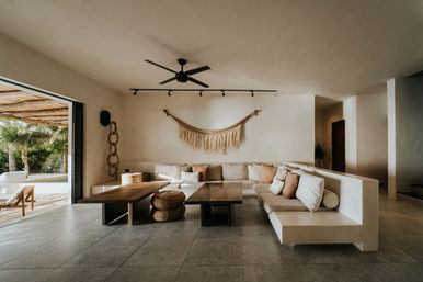 Sunlit coastal boho living room with U-shaped beige sectional, low wooden coffee tables, woven wall hanging, ceiling fan, and sliding doors to a palm-lined patio.