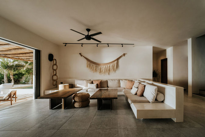 Sunlit coastal boho living room with U-shaped beige sectional, low wooden coffee tables, woven wall hanging, ceiling fan, and sliding doors to a palm-lined patio.