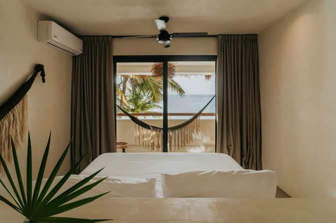Tropical beachfront bedroom with white bed and pillows facing sliding glass doors, balcony hammock under palm trees and a calm ocean view.