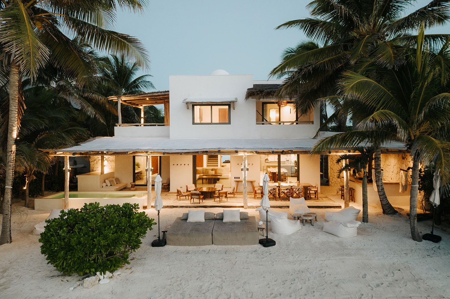 Tropical beachfront villa at dusk: modern white two-story house with open-air patio, dining and lounge seating on sandy shore framed by palm trees and warm interior lights.