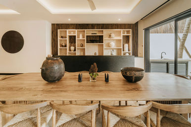 Sunlit modern-rustic dining area with a long wooden table and woven chairs, pottery and small plant centerpieces, black kitchen island with open wood shelving, and sliding glass doors to an outdoor terrace.