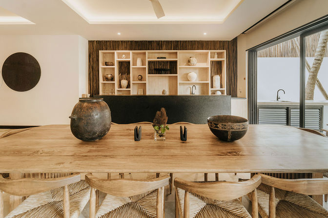 Sunlit modern-rustic dining area with a long wooden table and woven chairs, pottery and small plant centerpieces, black kitchen island with open wood shelving, and sliding glass doors to an outdoor terrace.