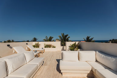 Sunny oceanfront rooftop terrace with white cushioned sofas and wooden lounge chairs, tropical palm trees and deep blue sky meeting the distant sea.