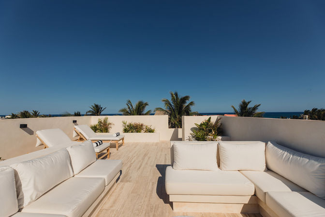 Sunny oceanfront rooftop terrace with white cushioned sofas and wooden lounge chairs, tropical palm trees and deep blue sky meeting the distant sea.