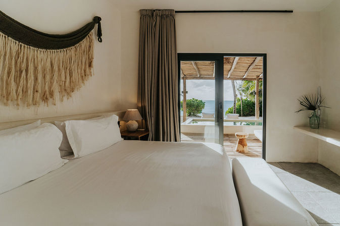 Sunlit beachfront bedroom with crisp white bedding, woven wall hanging and bedside lamp, sliding glass doors opening to a shaded patio and ocean view framed by tropical greenery
