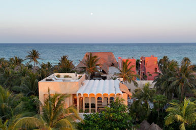 Aerial view of beachfront villas and palm trees along a tropical coastline at dusk — pastel pink and white buildings, calm blue ocean and moon on the horizon.