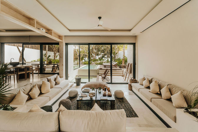 Airy coastal villa living room with built-in beige sofas and cushions, low rustic coffee table, sliding glass doors opening to a private pool, sun loungers and palm trees beyond