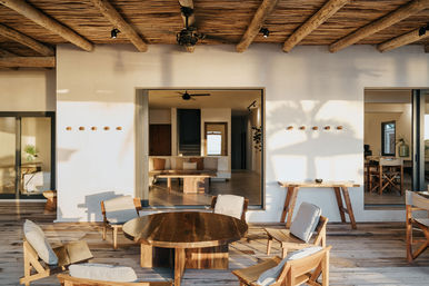 Sunlit outdoor patio with rustic wooden-beam ceiling and weathered deck, round wooden table surrounded by cushioned chairs, opening through large sliding doorways into a minimalist living room—warm, natural materials and relaxed coastal-style vibe.