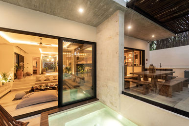 Modern resort-style villa with warm ambient lighting, open-plan living room and sunken seating seen through sliding glass doors, an illuminated private plunge pool and a covered outdoor wooden dining area.