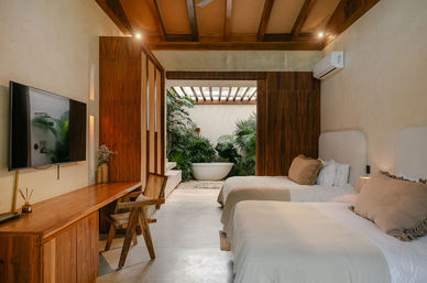 Cozy modern tropical villa bedroom with two twin beds, warm wood accents, wall-mounted TV and desk, opening to a lush private courtyard with a freestanding soaking tub.