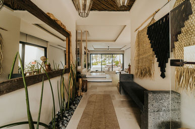 Beachside resort corridor with natural stone floors, woven wall hangings and macramé, long mirror and tall snake plants along a pebble planter, textured bench, and sliding glass doors opening to palm trees and ocean view