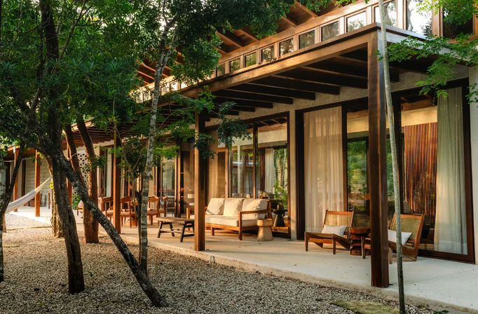 Sunlit modern wooden villa patio with cushioned sofa, wooden chairs and hammock under a covered terrace, surrounded by trees and a gravel courtyard