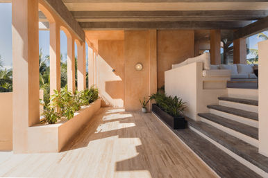 Sunlit tropical terrace with peach plaster arches, planters of greenery, steps up to a cushioned outdoor lounge and palm trees beyond