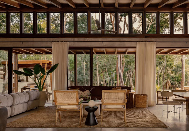 Cozy open-plan living room with high clerestory windows and floor-to-ceiling glass doors, exposed wooden beams, neutral sofa, rattan chairs on a jute rug, potted tropical plant and dining area opening to a lush forest view.