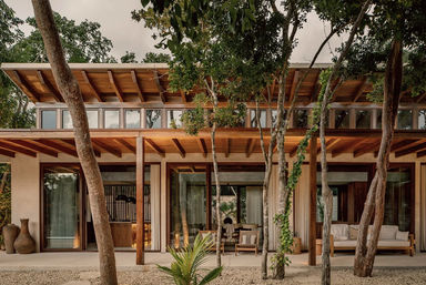 Modern tropical wood villa with wide overhangs and floor-to-ceiling glass doors, shaded outdoor lounge patio framed by trees and gravel courtyard