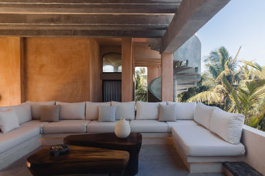 Open-air tropical balcony lounge with a large white sectional sofa, wooden coffee tables, terracotta plaster walls and a concrete spiral staircase overlooking palm trees.