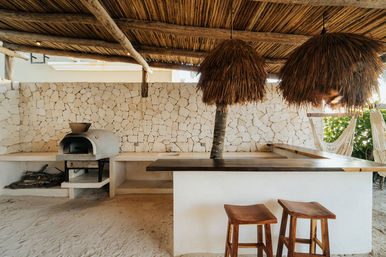 Tropical outdoor bar under a thatched palapa with stone wall, wood-fired oven, sandy floor, two wooden stools, hammock and a palm trunk through the counter
