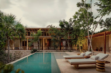 Tropical villa poolside at dusk with a turquoise rectangular pool, two cushioned wooden loungers and round side tables on a stone deck, framed by trees and warm-lit modern wooden architecture for a serene jungle retreat.