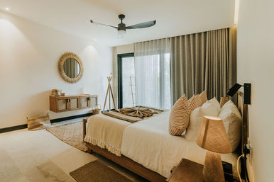Cozy modern boho bedroom with neutral tones: wooden platform bed dressed in cream linens and striped pillows, woven round mirror above a floating wooden console, ceiling fan, floor-to-ceiling curtains over a sliding glass door, textured rugs and warm ambient lighting.