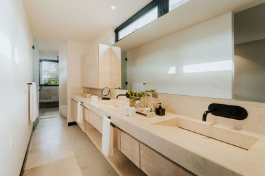 Sunlit modern bathroom with a long marble double vanity, integrated sinks, matte-black wall faucets, light wood cabinets, large mirror, white towels and a walk-in shower in the background.