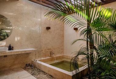Sunlit spa-like indoor courtyard bathroom with a square stone soaking tub filling from a wall spout, minimalist plaster walls, round mirror, floating sink shelf and lush palm plants.