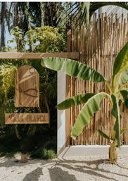 Sunlit tropical villa entrance with a carved wooden hanging sign, large banana leaves and palm fronds, and a bamboo privacy fence.