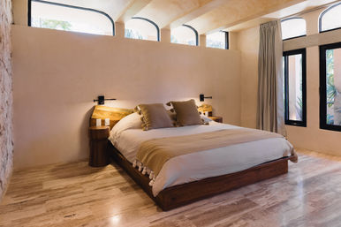 Cozy sunlit minimalist bedroom with low wooden platform bed, neutral linens, arched clerestory windows, and stone tile floor.